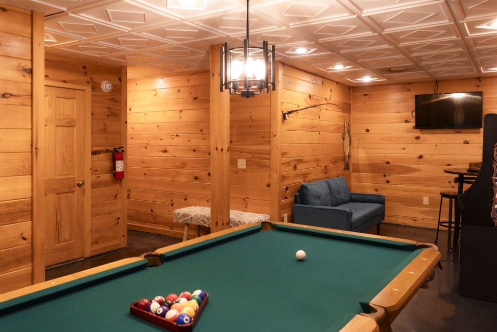 Game room of the Lincoln Logs Cabin with a pool table, sofa, small bench, arcade machine, tall table, and a television. The wall is decorated with a fishing stick